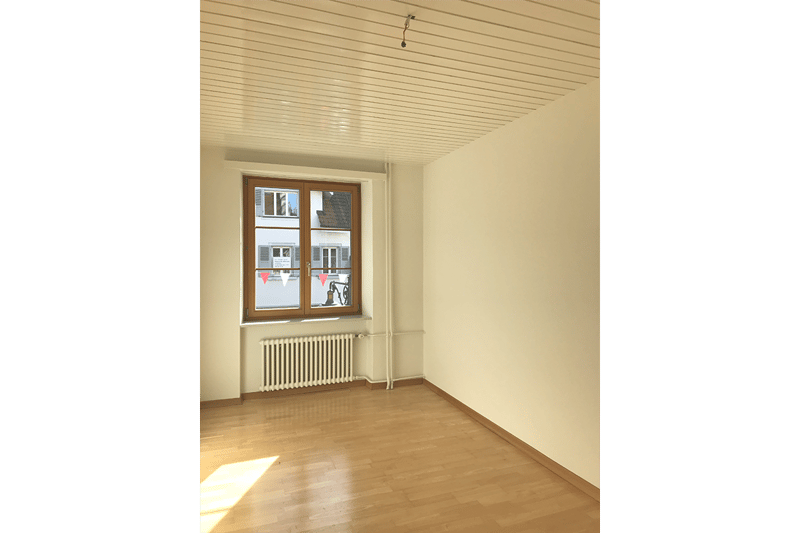 Empty room with wooden floor, large window, radiator on wall, ceiling beams.