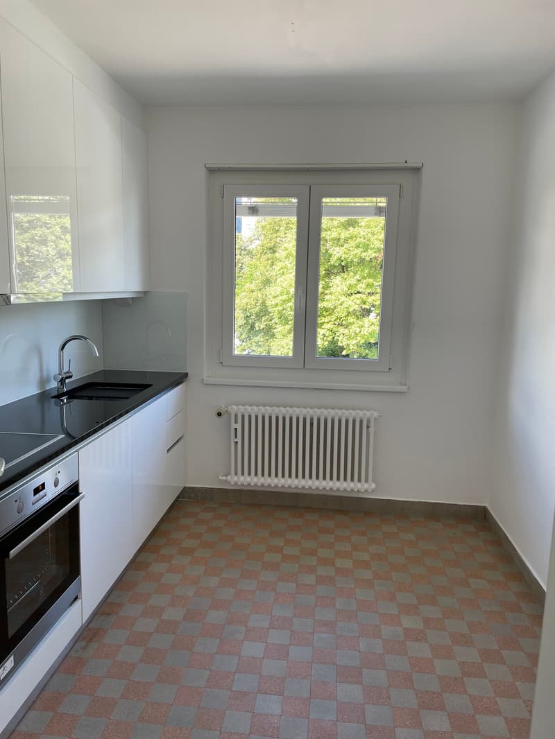 Empty kitchen with white cabinets, black countertop, white radiator, checkered floor, window with view of trees
