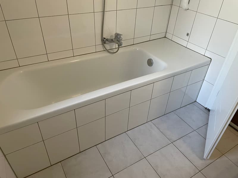 Bathroom with white tiles, bathtub, shower head, and open door.