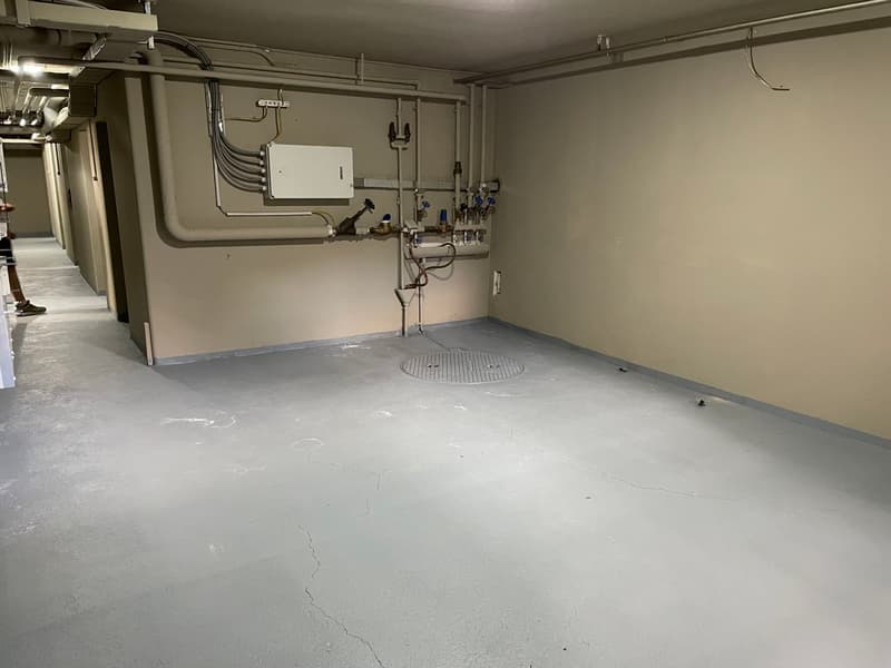 Empty room with concrete floor, pipes and valves on the wall, a manhole cover in the center, and a power outlet.