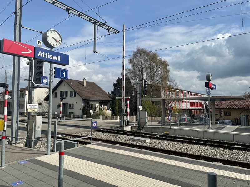 Railway station with platforms, tracks, and overhead wires. A blue sign with 'Attiswil' and a clock. Houses and a building in the background.