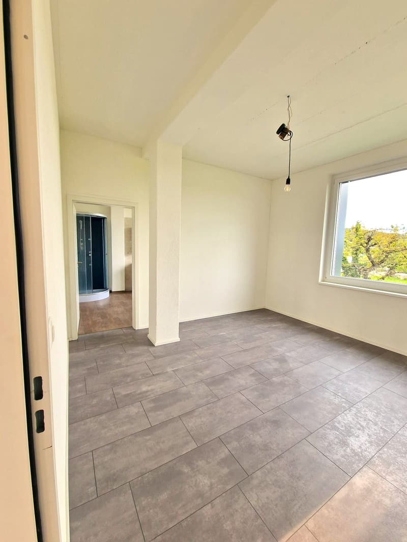 Empty room with tiled floor, white walls, large window, open door leading to another room with a shower