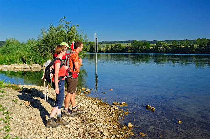 Three people standing on the shore of a lake, with a clear view of the water and surrounding greenery.