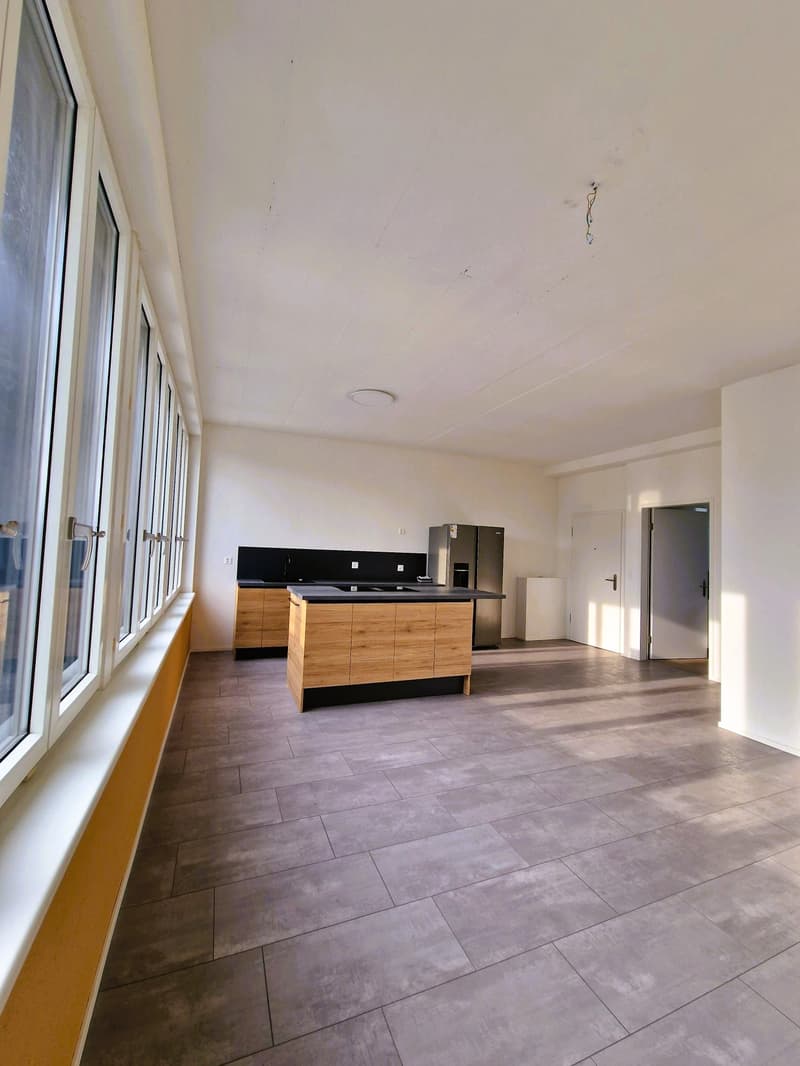 Empty kitchen with wooden cabinets, black countertops, stainless steel refrigerator, large windows, tiled floor