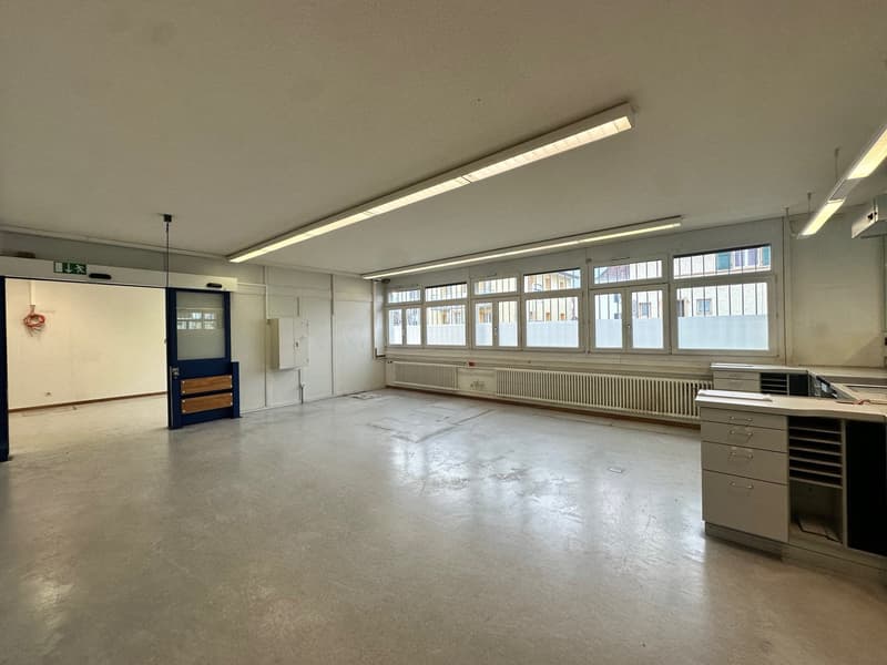 Spacious room with white walls, white ceiling, windows, and radiators. There are empty cabinets and an open door.