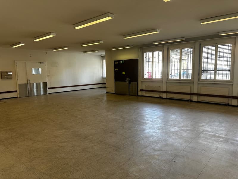 Large open space with white walls, white ceiling, square tiles, 2 doors, 4 windows with bars, fluorescent lighting