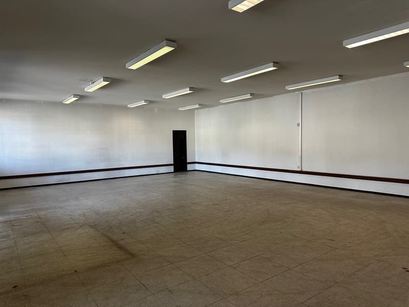 large open space, white walls, ceiling with strip lights, door, tiled floor
