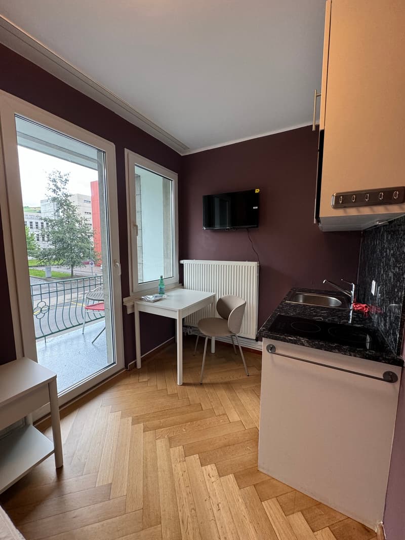 small room with a table, chair, and a television, a kitchen with a sink, stove, and a small refrigerator, a balcony with a view of the outside