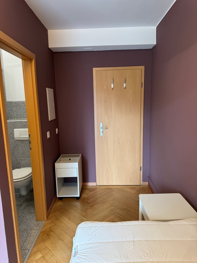 Bedroom with wooden floor, white walls, wooden door, small white table, bed, and a toilet in the adjacent room