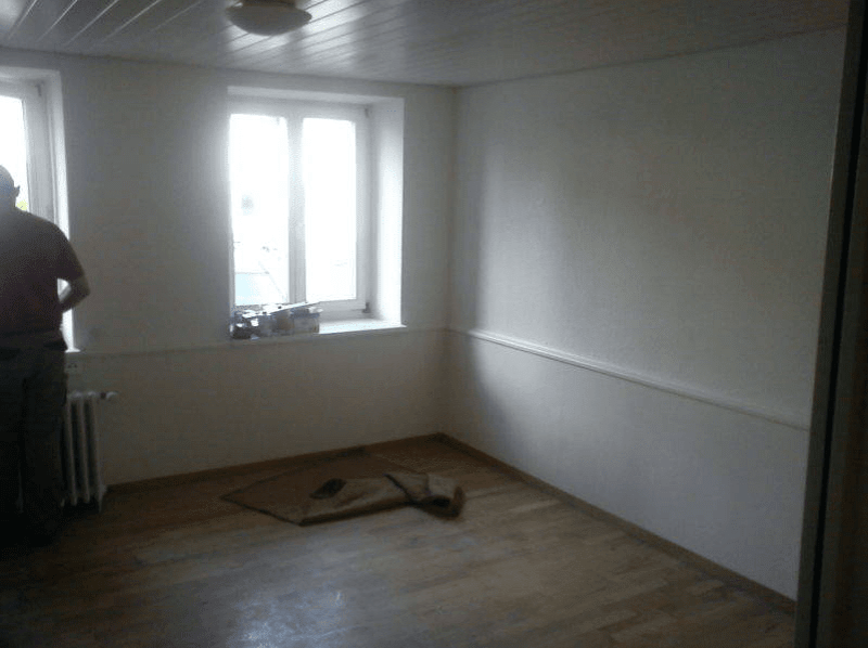 Empty room with white walls, wooden floor, two windows, radiator, items on window sill