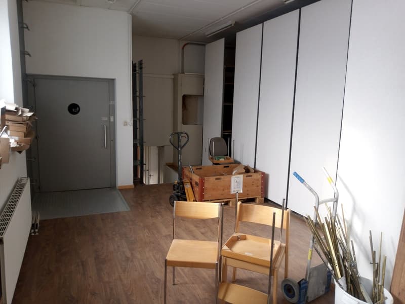 Empty room with wooden floor, white walls, gray door, wooden chairs, storage boxes, trolley, and radiator