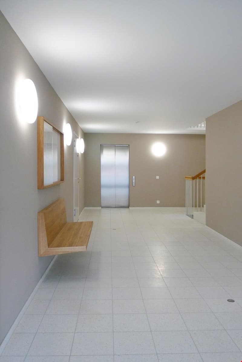 Empty hallway with white tiles, steel elevator, mounted mirror, ceiling lamps