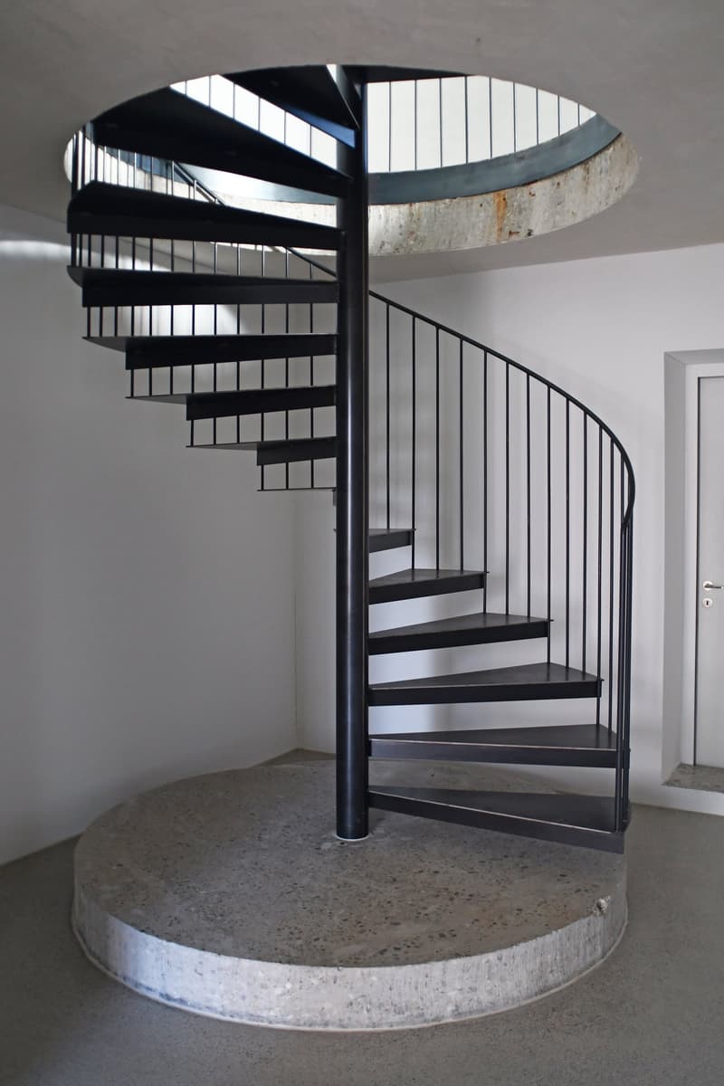 Black circular staircase with multiple steps, concrete base, glass railings.
