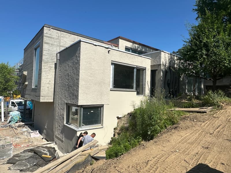 modern house under construction, beige and grey exterior, large windows, surrounded by trees, two people working on the side, construction materials on the ground