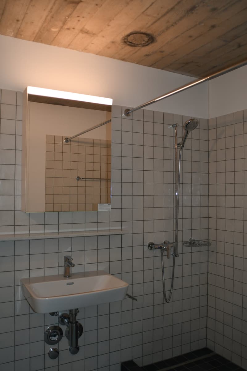 A bathroom with white tiles, a sink, a shower, and a mirror.