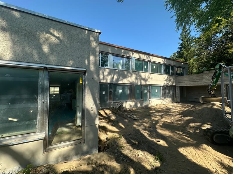 Single-story building with a modern design, multiple large windows, and exposed walls, under construction with a dirt area and construction equipment.