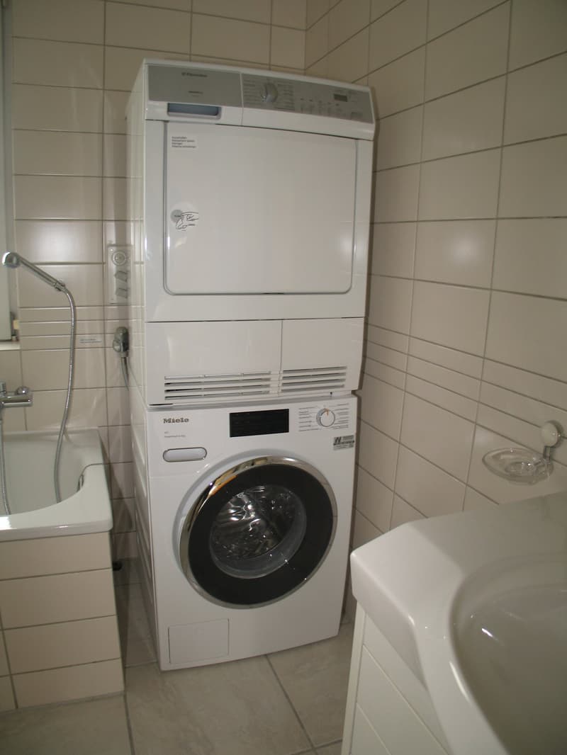 Bathroom with white tiled walls, stacked washer and dryer, bathtub, and sink.
