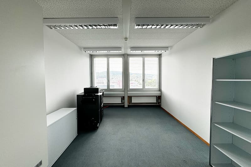 An empty room with white walls, gray floor, ceiling lights, a printer, and two windows with outside view.