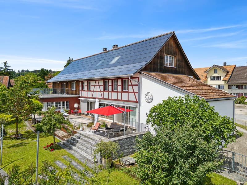 Detached house with solar panels, brown roof, large terrace with a red umbrella, garden, patio table, and chairs