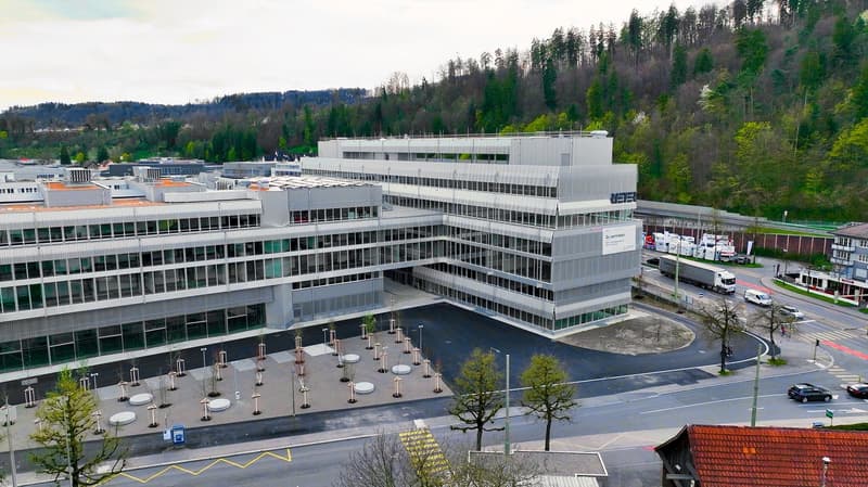 Large multi-story office building with a modern, glass and steel design. The building has a plaza in front with fountains and seating areas. The surrounding area includes trees and other buildings, suggesting a commercial or business district.