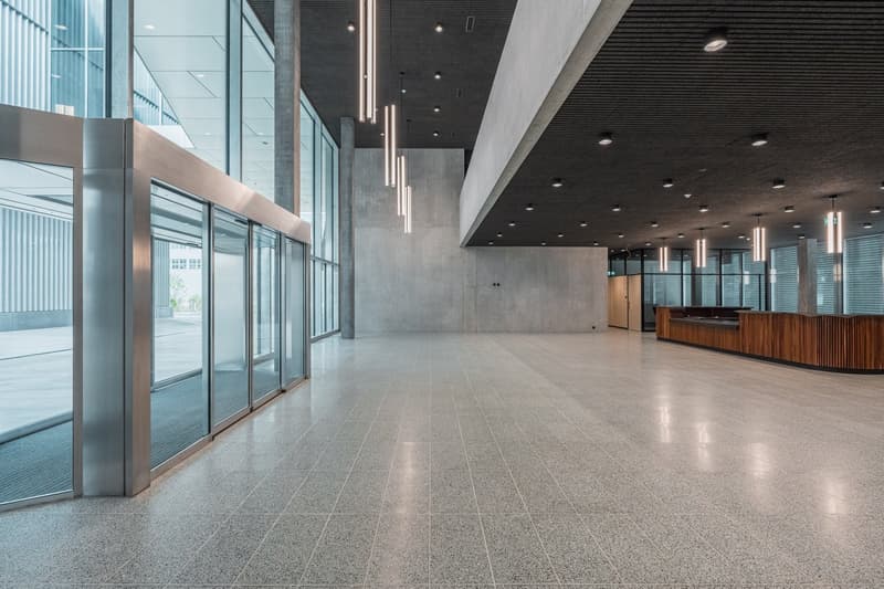 Large open space with concrete walls and floors, glass windows, and modern lighting fixtures. The space appears to be a lobby or entrance area of an office building.