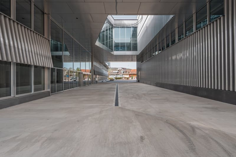 The image shows a modern commercial building with a large open space in the center. The building has a sleek, minimalist design with glass walls and a high ceiling. The floor is made of concrete, and there are no visible features such as balconies, garage