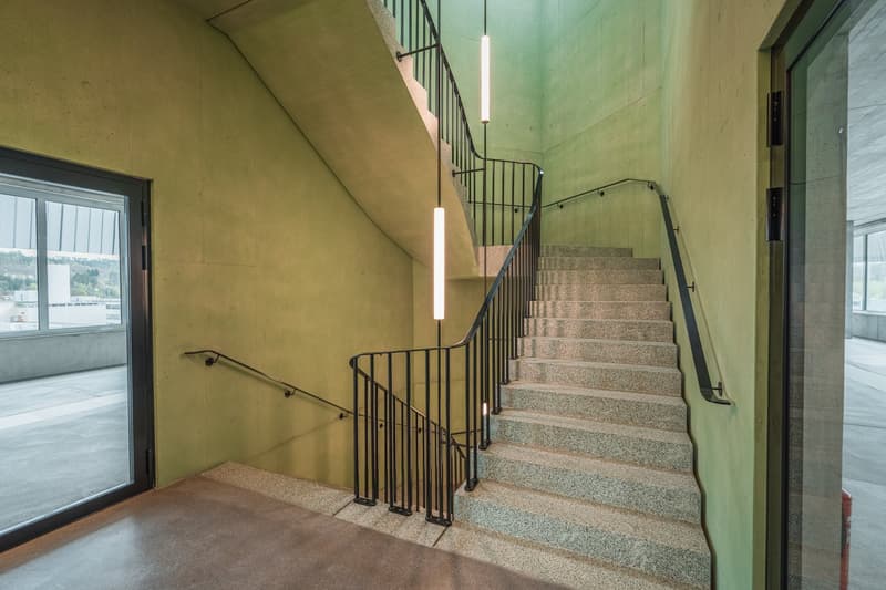 Concrete staircase with metal railings, leading up to a higher level. The walls are painted in a light green color, and there are large windows providing natural light.