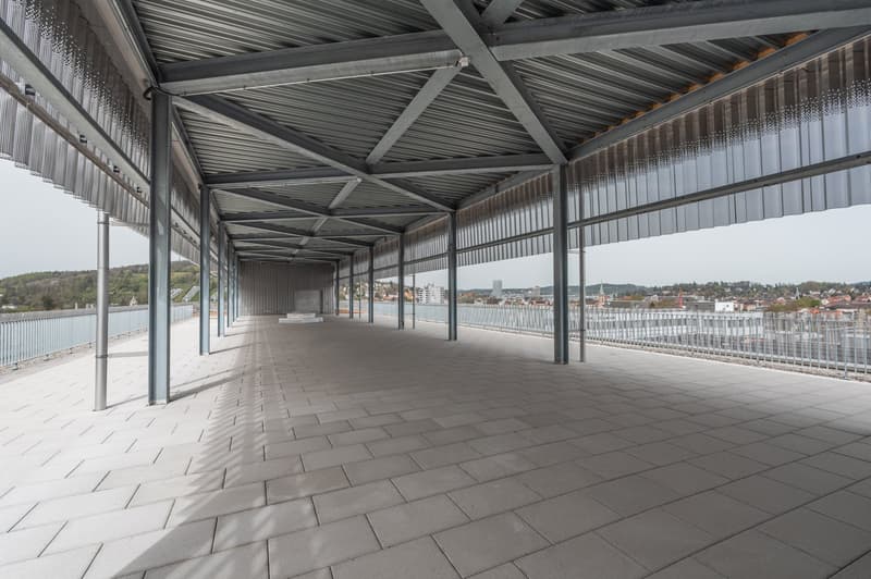 Large covered outdoor area with a metal roof structure, concrete floor tiles, and railings along the sides. The area overlooks a cityscape in the distance.