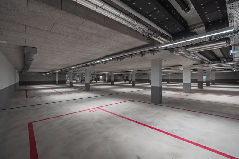 Large indoor parking garage with concrete floors, red parking lines, and support columns. The ceiling has exposed ventilation ducts and lighting fixtures.