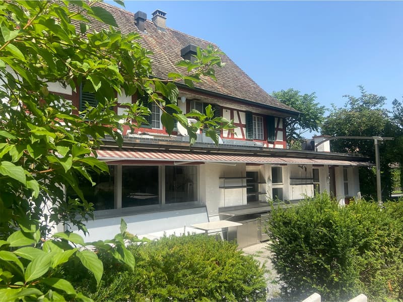 Two-story house, brown roof, white and red walls, red and white awning, kitchen area with windows, shelves, and a table, surrounded by trees and bushes.