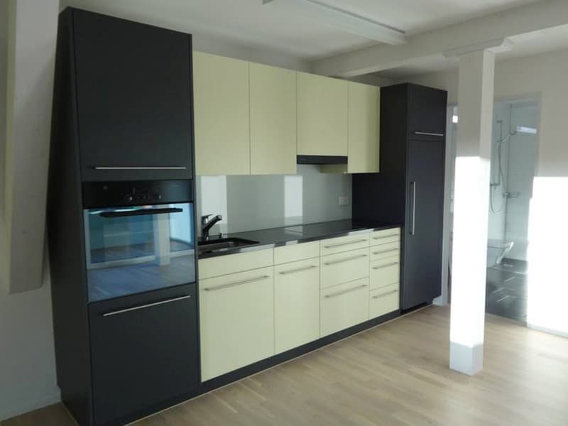 Modern kitchen with black and beige cabinets, wooden floor, sink with faucet, oven, dishwasher, countertop, and a view to the bathroom