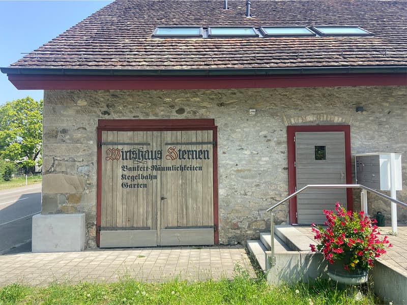 Stone exterior, double wooden doors, two windows above doors, staircase on the right, small handrail on the left, sign, small flowerpot on the ground in front, mailbox on the right side