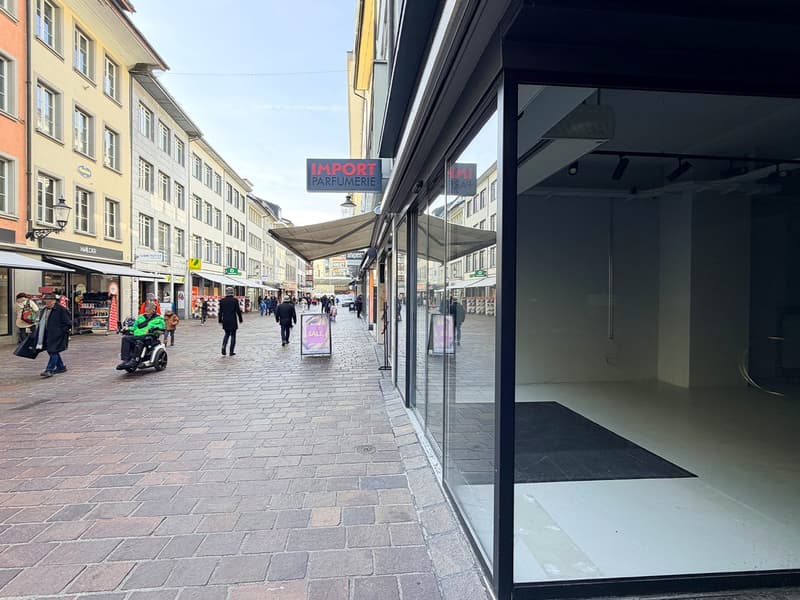 Outdoor view of a city street with buildings, people walking, an empty storefront, and a sign reading 'Import Parfümerie'.