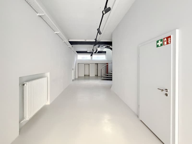 wide white walls, white floor, radiator, door on the right, emergency sign, corridor with staircase, windows