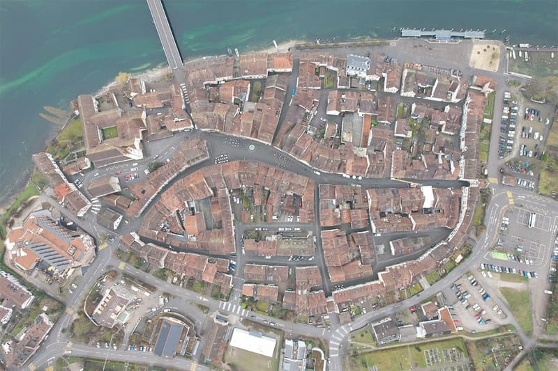 Aerial view of a town featuring various residential buildings with brown roofs, a central roundabout, multiple parking areas, and roads surrounded by green spaces and water.