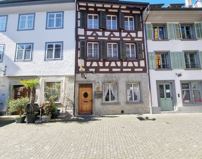 3-story building with a wooden door, many windows, and stone wall, 2 potted plants in front, brick pathway