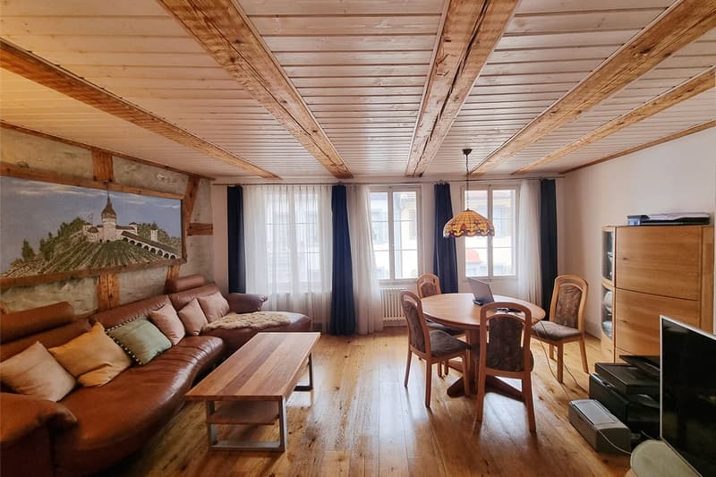 large couch with pillows, coffee table, dining table and chairs, wooden beam ceiling, painting, white windows with curtains, laptop on table