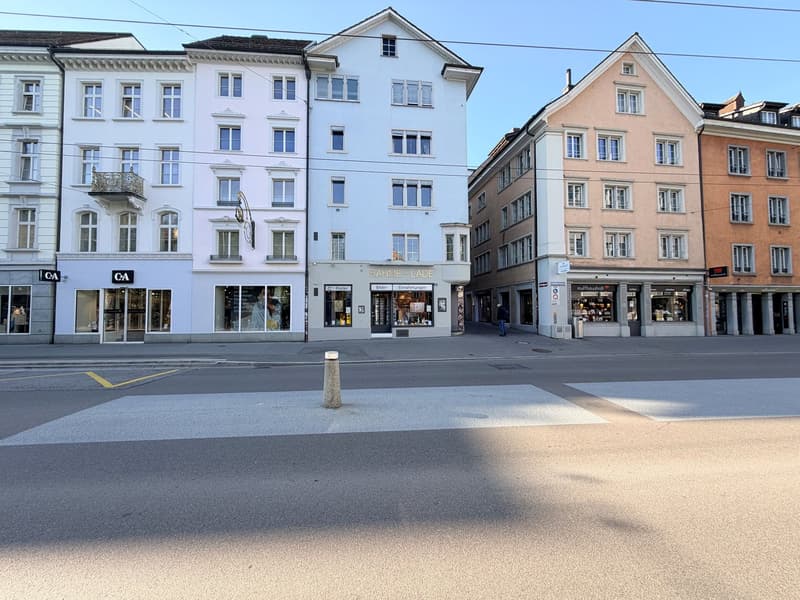 A street with several buildings, one of which has a sign that reads 'RAHME-LADE'. The buildings are of different colors and have many windows. There is a person walking on the sidewalk in front of the buildings.