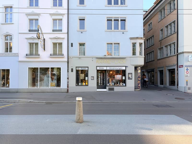 A street view of a building with glass windows and a sign that reads 'RAHME - LADE'. There is a person riding a bicycle on the sidewalk. The building has a glass door and a glass window with a display of items inside. There is a pole in the middle of the