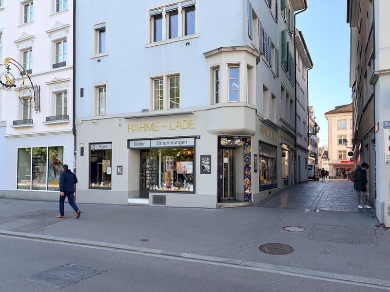 A three-story building with a shop on the ground floor, featuring a sign that reads 'RAHME-LADE'. The shop has large glass windows displaying various items. The building has a modern architectural design with white walls and green window shutters. There a