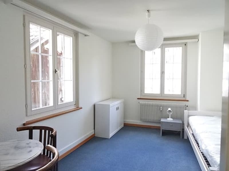 A small room with a bed, a desk, a chair, a cabinet, and a lamp. The room has white walls, a blue carpet, and two large windows with wooden frames. The bed is placed against the wall, and the desk is placed near the window. The room has a radiator and a l