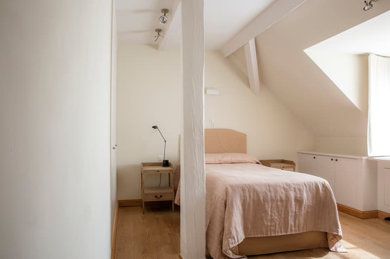 Spacious bedroom with slanted ceiling, hardwood floors, white walls, and a large bed with a pink linen bedspread. There is a wooden nightstand with a lamp and a cabinet or dresser against the wall.