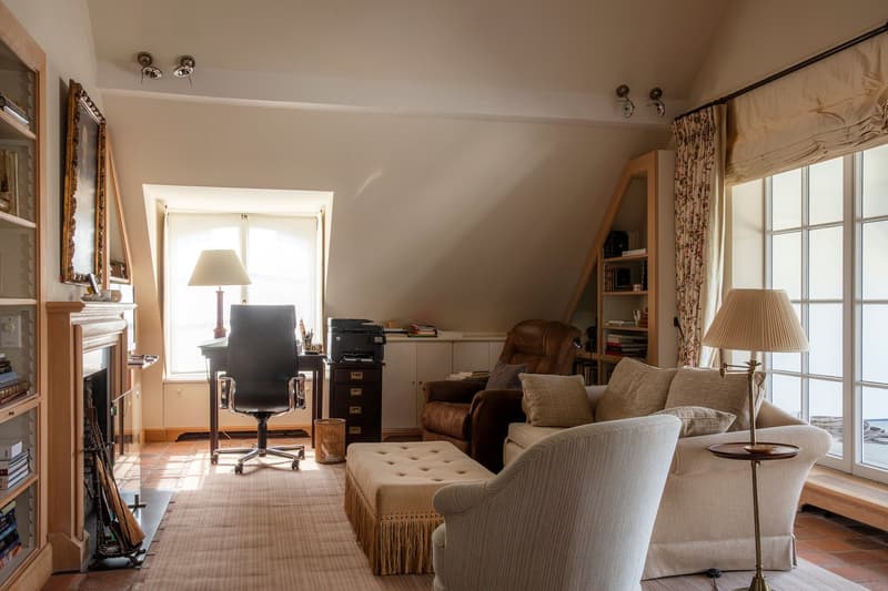Cozy living room with large windows, beige sofa, armchair, coffee table, desk with chair, bookshelves, and curtains. The room has a warm, inviting atmosphere.
