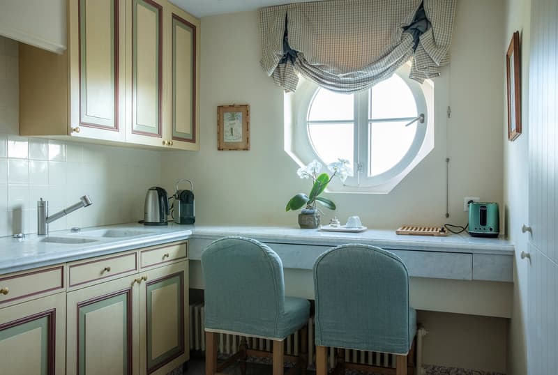 Spacious kitchen with wooden cabinets, marble countertop, round window, dining area with upholstered chairs, kitchen appliances including a dishwasher, and a decorative plant on the counter.