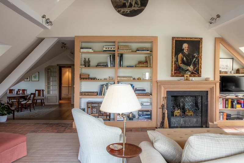 Cozy living room with a fireplace, built-in wooden bookshelves, a white armchair, and a framed painting on the wall. The room also features a dining area with a wooden table and chairs.