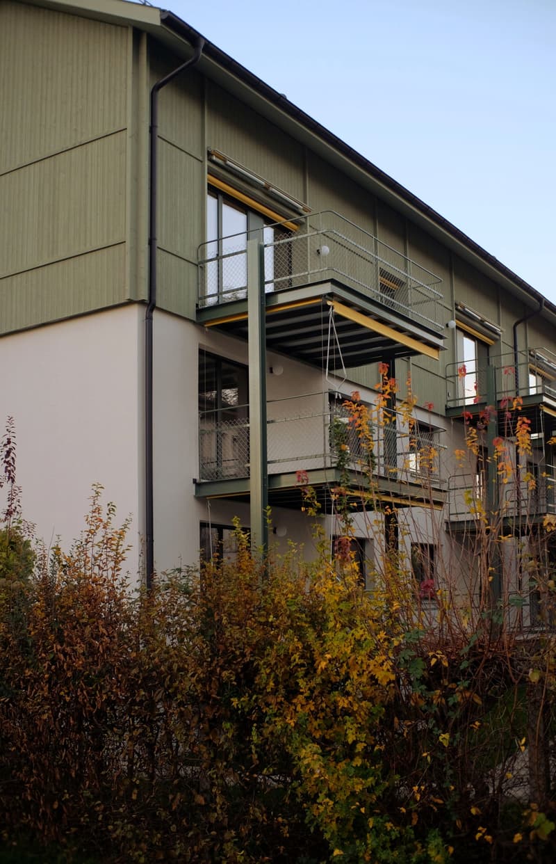 Building with balconies and railings, green and white exterior, plants, and bushes in front.