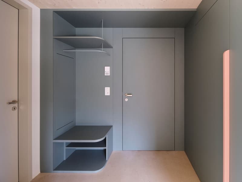 Minimalist entryway with gray walls, integrated shelving, and metal door with silver handle and lock. A hanging rod and a bench-like shelf are attached to the wall.