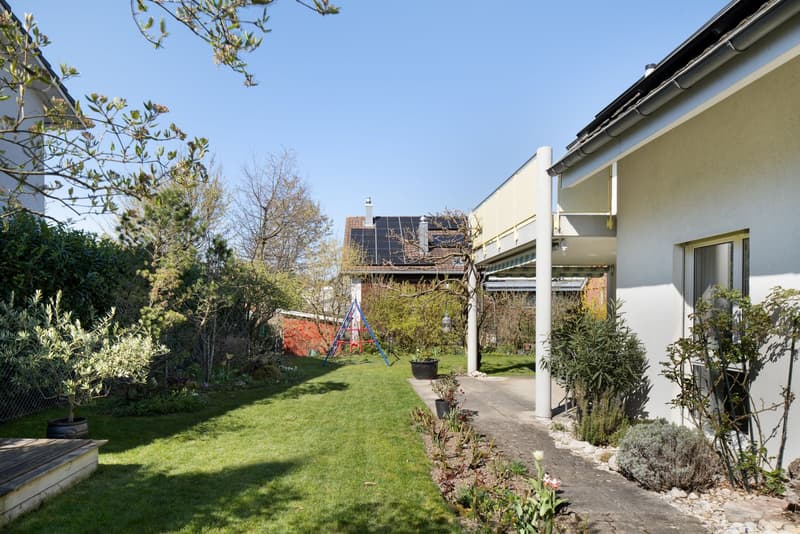 House with solar panels, garden with lawn, plants, and trees, pathway, swing, and a wooden deck.