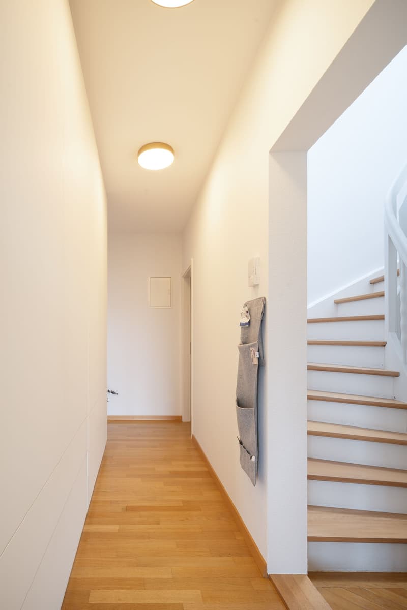 Empty hallway with wooden floor, white walls, ceiling light, and a staircase.