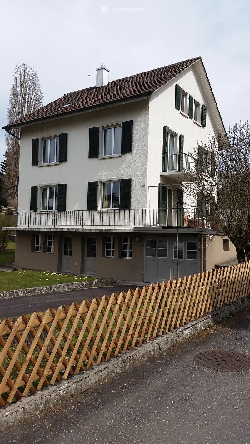 Two story house, brown roof, balcony, fenced garden, garages, white exterior, green windows, chimney, grass lawn, wooden fence